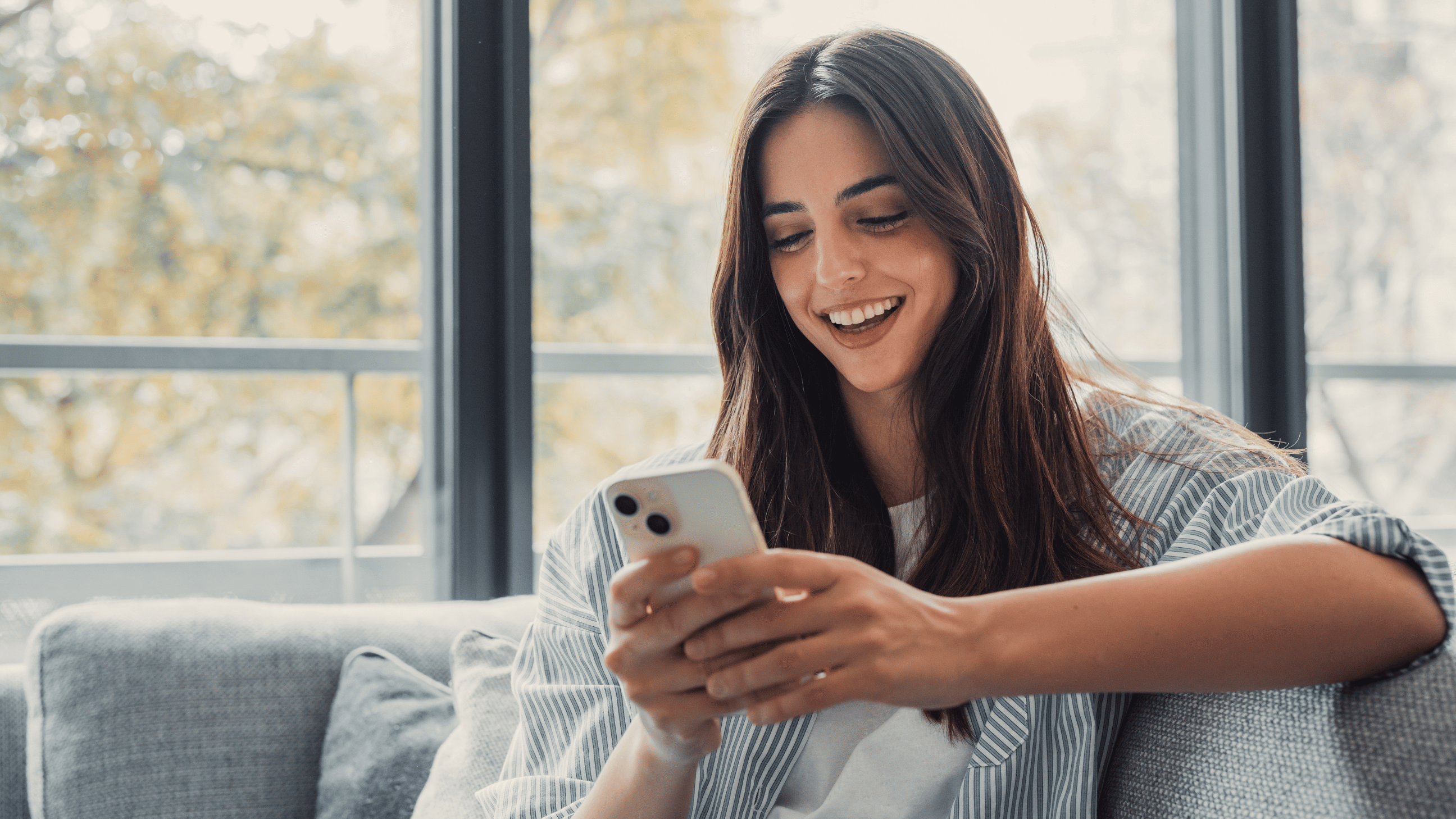 A brunette woman smiles on the couch as she looks at her phone - Aven Hospitality