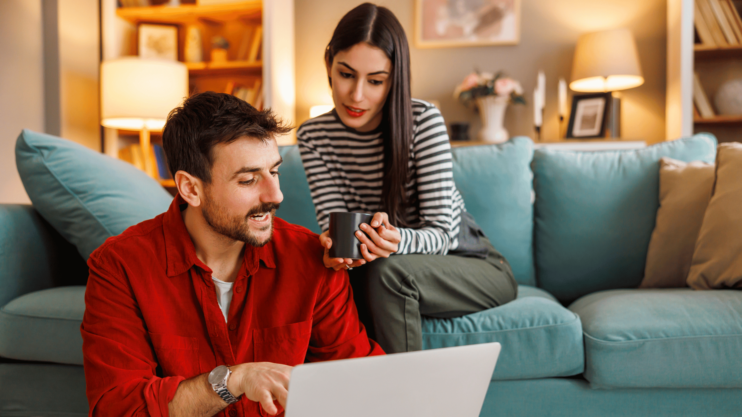 A man and woman sit in a living room, looking at a computer and phone while smiling - Aven Hospitality